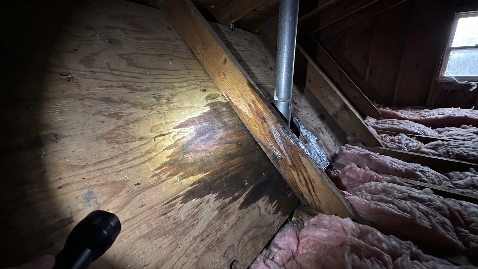 Attic view of a dark water stain spreading on roof decking and a wet rafter beside a plumbing vent pipe